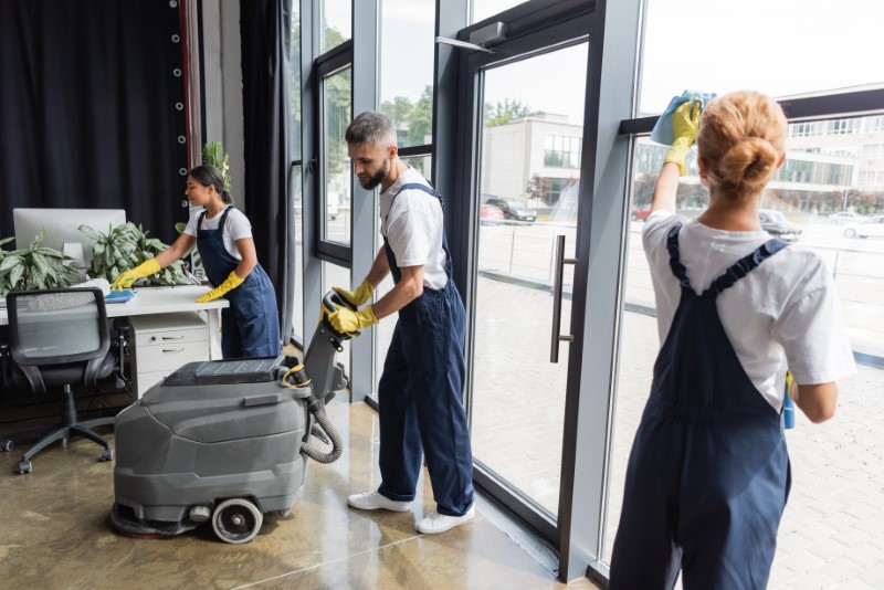 man with electrical floor scrubber machine near interracial women cleaning office.